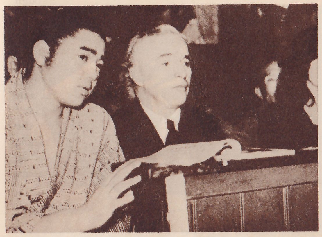 Close photograph of two men seated side by side. At the left a young man in a robe patterned with short dark dashes, with his hair swept back, looks to one side, one hand open in front of him at the lower edge. Next to him an older man with pale hair, in a dark jacket, white collar and dark tie, looks off in the same direction, one hand resting on the top of a dark paneled rail in front of them. Other people are only partly seen in the dark at the right.