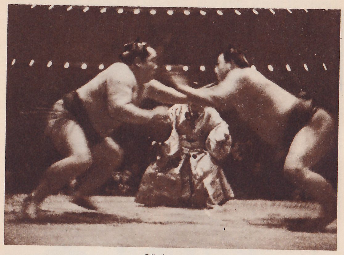 Photograph, blurred by motion, of two heavy bare men rushing at each other from the left and right with their arms stretched toward each other, each with a dark belt. Between and behind them a man in a pale patterned robe crouches low near the floor. A string of small white lights runs across the dark background in two rows.