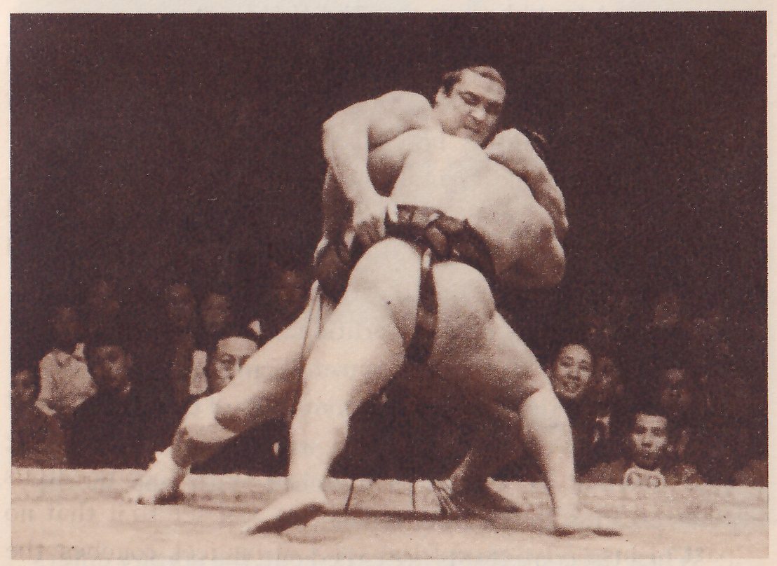 Photograph of two heavy men gripping each other at the center of a platform. One stands behind the other with his head up and a hand at the other man's belt at the middle of his back, while the nearer man, seen from behind with his legs spread wide and a dark belt showing, is bent forward. A row of spectators sits behind the edge of the platform.