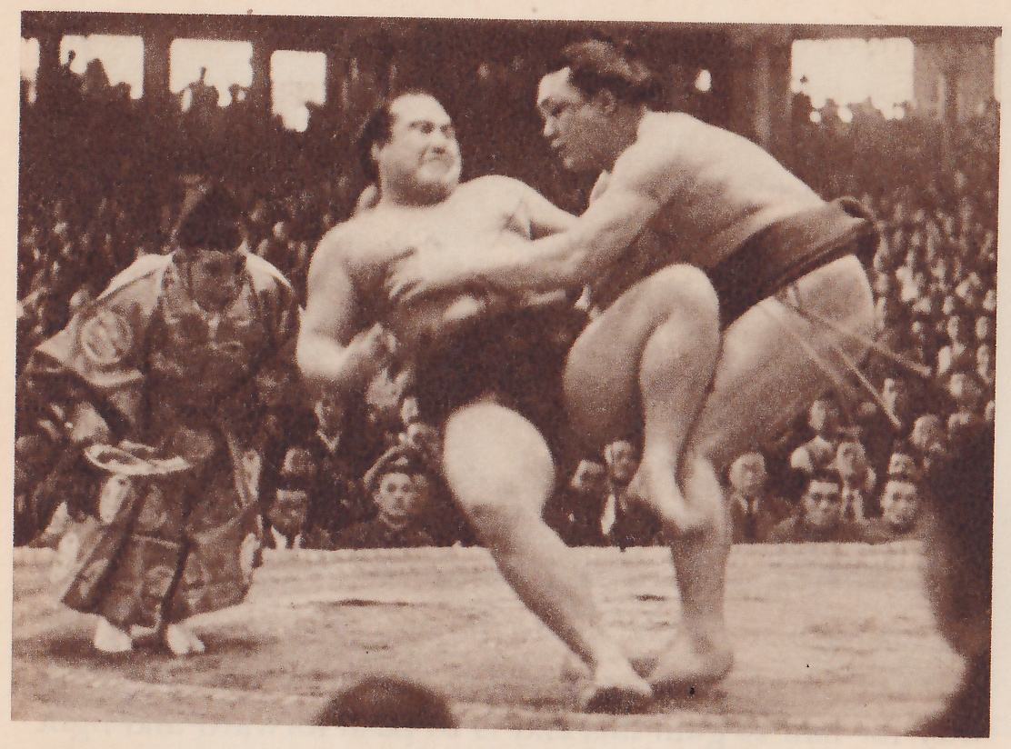 Photograph of two heavy bare men at the center of a platform. The man at the right leans in with an arm stretched against the other's chest and his near leg lifted, bent at the knee. The man at the left leans back with his mouth open and his face tense. At the left a man in a patterned robe and dark cap crouches, bent forward, holding a flat rounded object. Spectators fill the hall behind.
