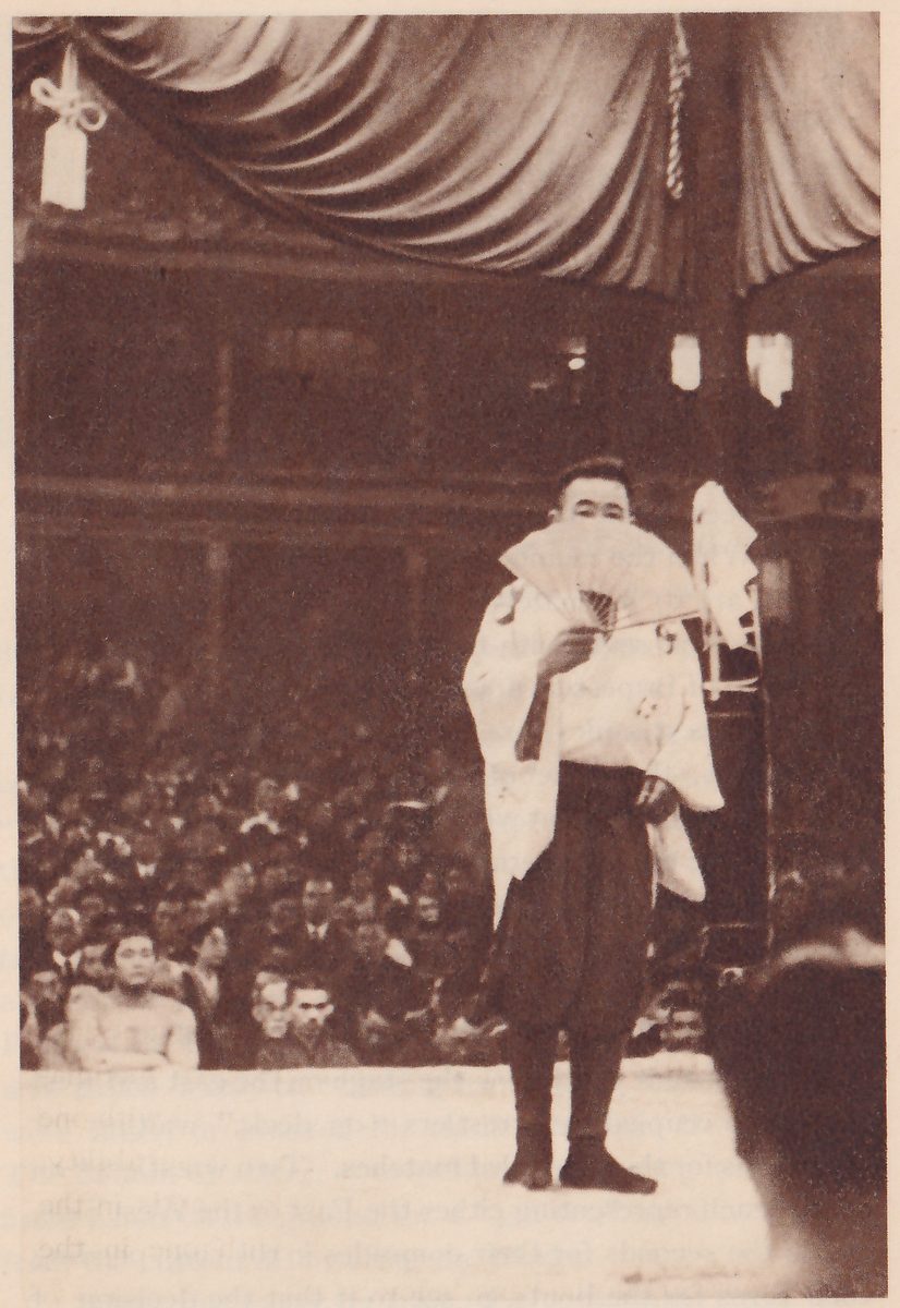 Tall photograph of a man standing alone on a pale flat platform, holding an open folding fan in front of his lower face. He wears a pale loose robe over dark wide trousers and dark footwear. A draped curtain with a hanging tassel fills the top, tiers of spectators fill the background, and a dark blurred head fills the lower right corner.
