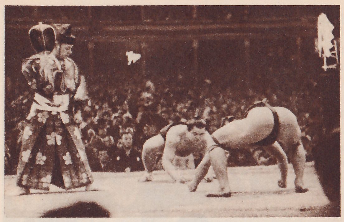 Photograph of a platform with two heavy men crouched facing each other at the center right, the one at the left with his head up and a hand on the floor, the other bent over with his back to the camera. At the left stands a man in a long robe with a large pale flower pattern and a black cap, with a white sash at his waist. A pale post stands at the upper right, and a crowd fills the hall behind.