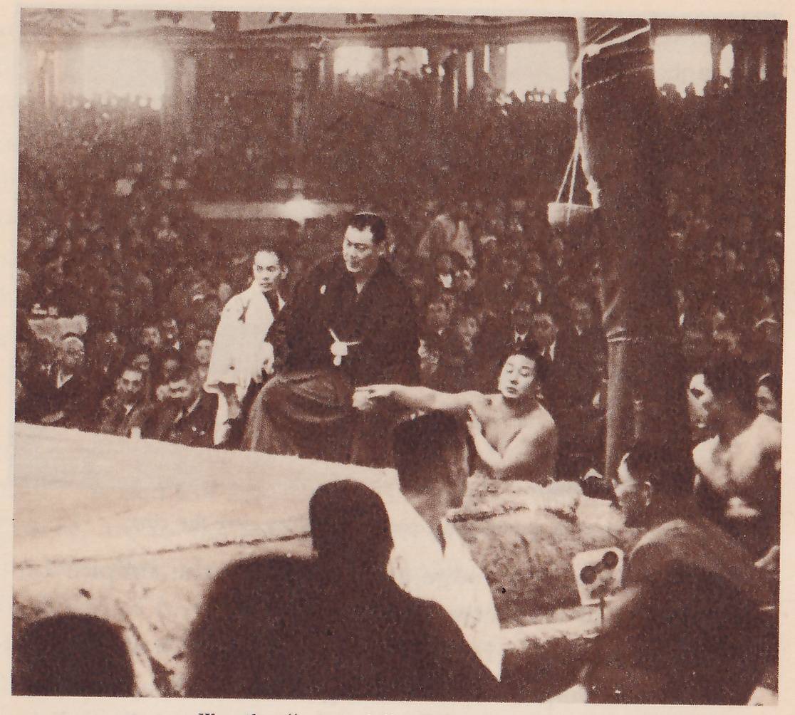 Photograph across the edge of a raised platform. A man in a dark robe sits at the center behind the edge, with a man in a white jacket standing behind him at the left. A bare-chested man sits in front of him at the right and stretches one arm out toward the seated man. A second bare-shouldered man is at the far right beside a thick post tied with rope. In the foreground the dark heads of men and a man in a white jacket look toward the platform, and a crowd fills the hall behind.