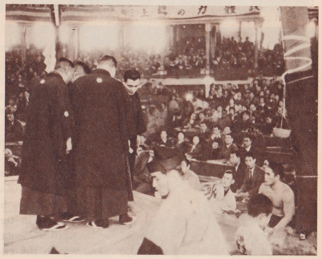Photograph of several men in long dark coats with a round pale crest on each coat, standing close together at the left of a platform with their heads bent toward one another. In the foreground a man in pale clothes crouches, and a bare-shouldered man kneels at the lower right. Tiers of spectators fill the hall behind, a banner of Japanese characters runs across the top, and a thick post with a spiral of white streamer stands at the upper right.