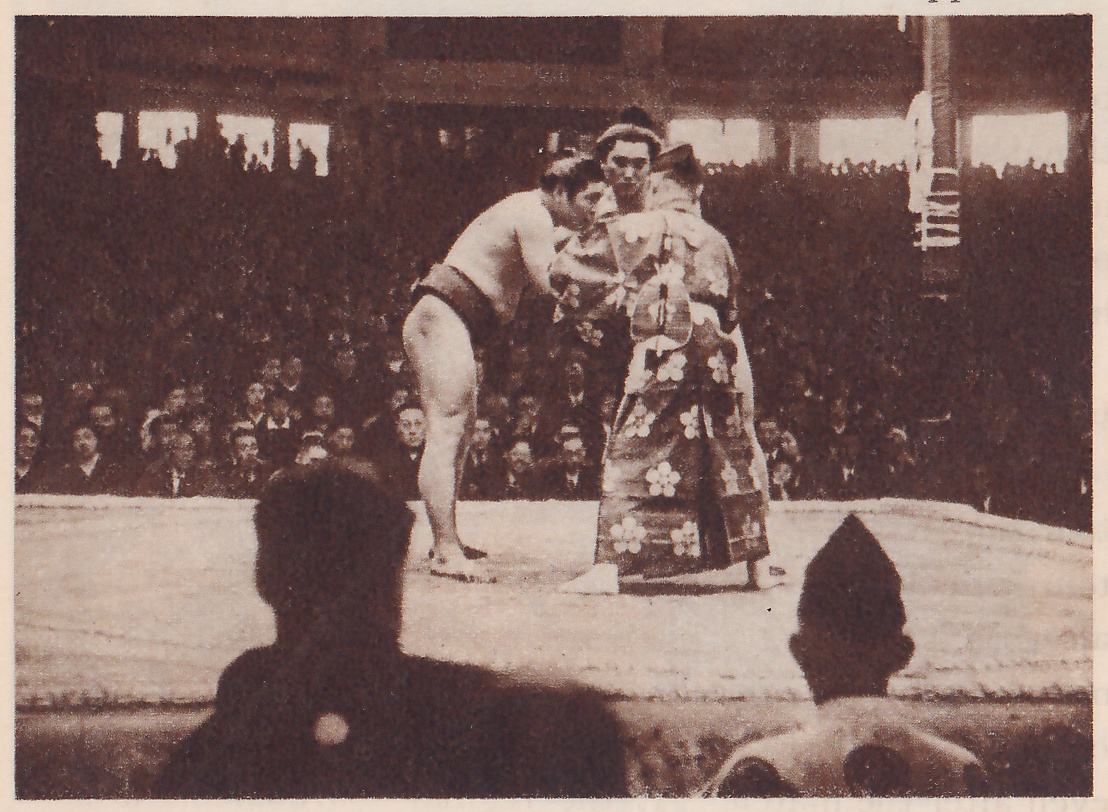 Photograph across the edge of a platform. At the left a bare-chested man in a dark belt leans forward with his arms out toward a man who stands at the right in a long robe with a large pale flower pattern. Behind them another man with a topknot looks on. Rows of spectators fill the background. In the foreground the dark head and shoulders of a man are at the left, and a head in a pointed black cap is at the lower right.