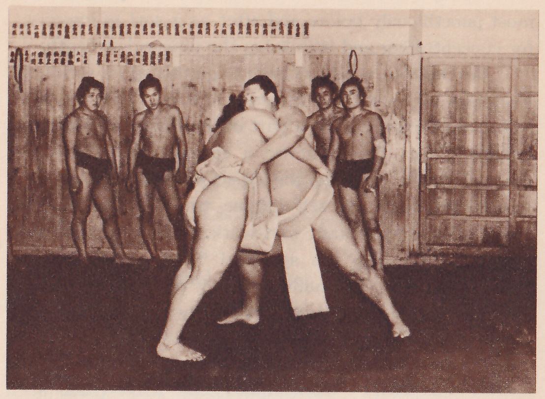 Photograph indoors of two heavy men locked in a close hold at the center, each with his arms around the other and a long pale cloth hanging down between them from the belt. Four slimmer bare-chested men in dark briefs stand against a wooden board wall behind, two at the left and two at the right, watching. A paneled sliding door is at the right and a rope hangs on the wall. The floor is dark and bare.
