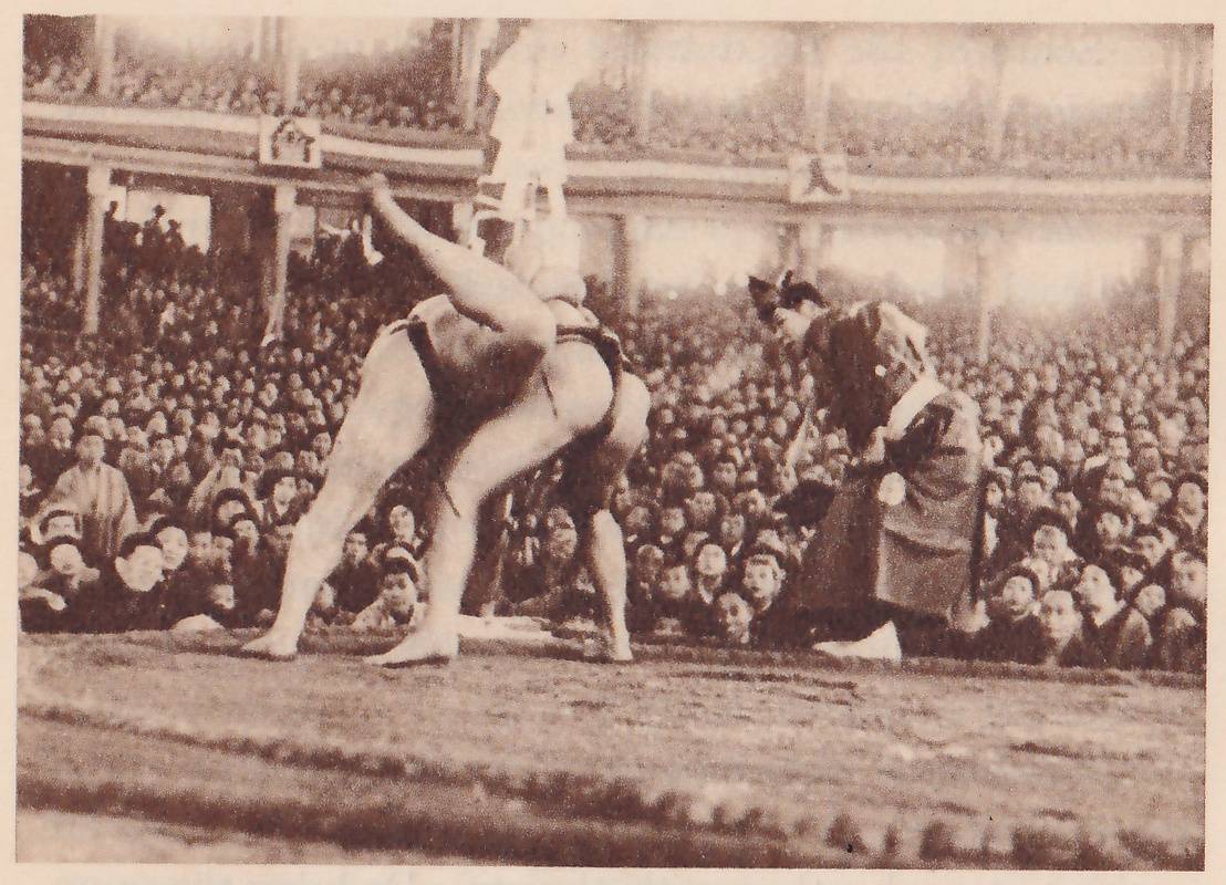 Photograph of two heavy men locked together at the center of a platform, bent forward, one with a leg raised high behind him toward the upper left and the other with his feet spread on the floor. At the right a man in a long robe and a black cap bends forward to watch. A pale column with white streamers stands behind them, and spectators fill the tiers and the galleries, which carry small framed pictures.