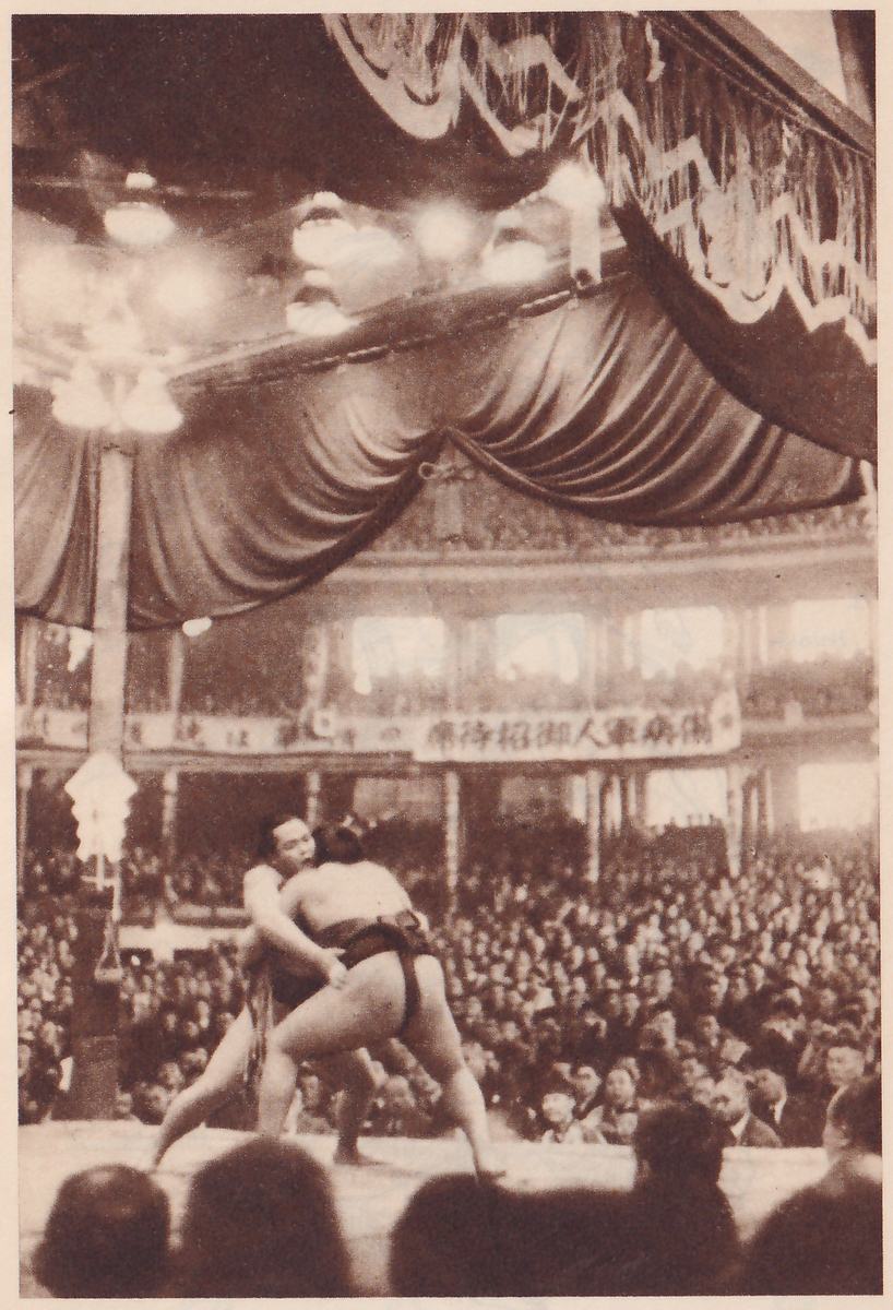 Tall photograph of a hall. Dark gathered curtains and a heavy garland with a ruffled, patterned edge hang across the upper part, with glowing lamps among them and a pale post at the left. A banner of Japanese characters runs along the gallery railing. At the center two heavy men clasp each other on a platform, one with his arm around the other, and a crowd fills the hall around them, with the dark heads of spectators at the bottom edge.