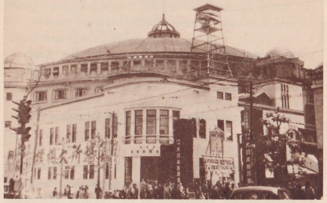 Photograph of a large building seen from a street corner. A wide low dome with a small domed cap at its top rises behind a pale curved-front lower building, and a tall open scaffold tower stands against the dome at the right. The lower building has tall windows grouped in a bay at the center, a dark doorway below it, and signs and hanging banners with writing at street level. Small figures of people stand along the street, a traffic signal is at the left, a car shows at the lower right, and trees stand at the right.