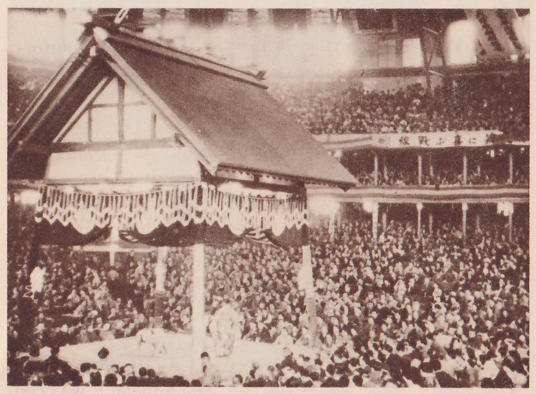 Photograph looking down into a very crowded hall. At the left a large gabled roof with a wooden frame under its gable and a valance of white scalloped lace-like trim over dark swags stands on posts above a pale platform, on which a few small figures show at the lower center. Spectators fill the floor in dense rows and the galleries at the right, and a banner of Japanese characters runs along the gallery railing. Bright haze spreads across the top of the picture.