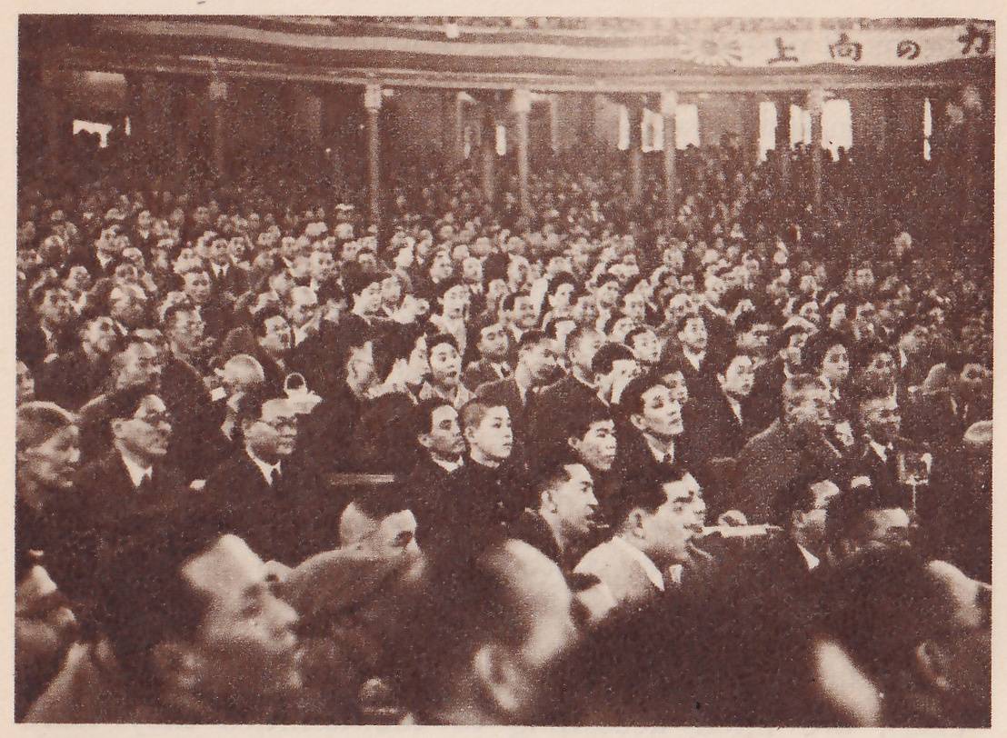 Photograph of a closely packed crowd of spectators seen from among them, most of the men in dark suits and ties, some in caps, with several faces turned toward the same point at the right. Columns and balconies show at the back, and a banner of Japanese characters runs along the top edge. Heads and shoulders fill the lower foreground.
