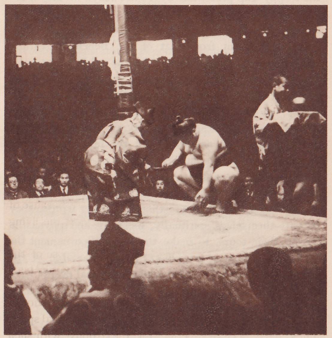 Dim photograph of the edge of a pale platform. A bare-chested man in a dark belt squats low at the center with one hand near the floor and the other stretched toward a man in a light patterned robe who bends toward him from the left. At the right a man in a pale robe stands behind a draped table or stand with a small pale object on its top. A post wrapped with paper strips stands at the upper left. Spectators sit in rows behind, and the dark heads of men, one in a pointed black cap, fill the foreground.