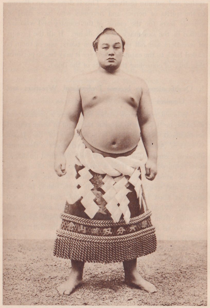 Photograph of a heavy man standing facing the camera with his feet apart and his arms at his sides, bare to the waist, with a topknot. A thick twisted white rope is wound around his middle, and an apron with white zigzag paper strips hanging over it covers the front of his dark belt. Below that is a wide band with lettering in Japanese characters between two thick cords, and a deep fringe of twisted cords hangs at the hem. He stands on bare gritty ground against a plain backdrop.