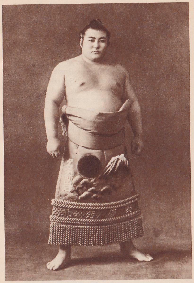 Photograph of a heavy man standing facing the camera with his arms at his sides, bare to the waist, with a topknot. A wide folded belt of pale cloth is at his waist, and an apron hangs below it decorated with a dark disc, a peaked shape with white streaks and rounded shapes below them, with Japanese characters in a vertical line at the left edge. Below that are a band with lettering in Japanese characters between two thick cords and a deep fringe of twisted cords at the hem. He stands on bare ground against a plain mottled backdrop.