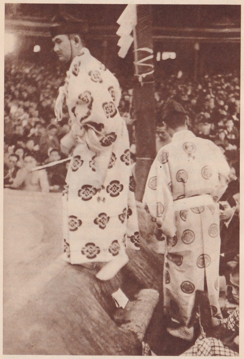 Photograph of two men in long pale robes patterned with large dark round crests, each wearing a black cap, at the edge of a raised platform. The man at the left is seen from the side with his head turned to the left, and a thin pale rod points out to the left at his waist. The man at the right is seen from behind. A thick dark post with white paper strips and a spiral cord at its top stands between them, a crowd fills the hall behind, and a plaid-patterned cloth and a seated man show at the lower right.