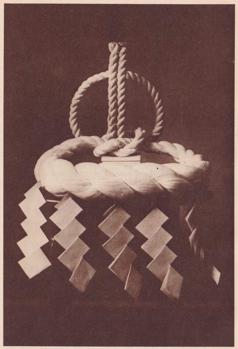 Photograph against a dark background of a thick pale twisted rope coiled into a wide ring, with a knot of the rope at the top center and an upright loop of thinner twisted rope standing above it. Chains of folded white paper strips in a zigzag hang from the thick ring, four of them across the front and more at the sides.