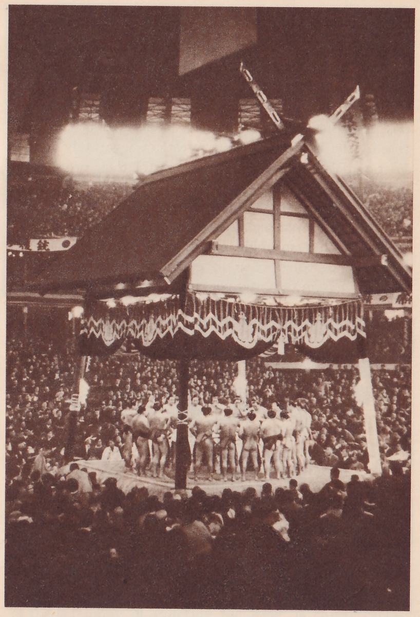 Photograph looking down into a very crowded hall. A large gabled roof with a dark valance edged with white zigzag trim stands over a platform at the center, and a group of men in belts stand side by side in a row on the platform, around a post. Spectators fill the floor and the galleries behind the roof, and the dark heads of the crowd fill the foreground. Bright haze spreads across the upper part.