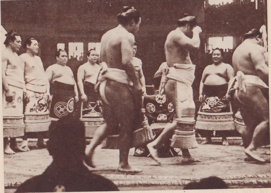 Photograph of a platform with men in aprons. In the foreground two bare-chested men in pale belt cloths walk across the center, one seen from the side and the other with an arm raised to his head, and a third man is partly seen at the right edge. Behind them a row of men stand in aprons marked with large round crests and with deep fringes at the hem, some of them smiling. The dark head and shoulders of a spectator fill the lower left.