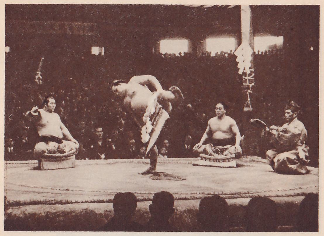 Photograph of a broad flat platform. A heavy man at the center bends forward at the waist on one leg, one arm lifted behind him, in an apron with a hanging pale panel. A bare-chested man sits on a low bundle at the left with one hand raised, another sits on a low bundle at the right of center, and at the far right a man in a patterned robe and black cap kneels holding a flat object. A pale post with paper strips stands at the upper right, a band of writing shows along the upper left, and the dark heads of spectators line the bottom.