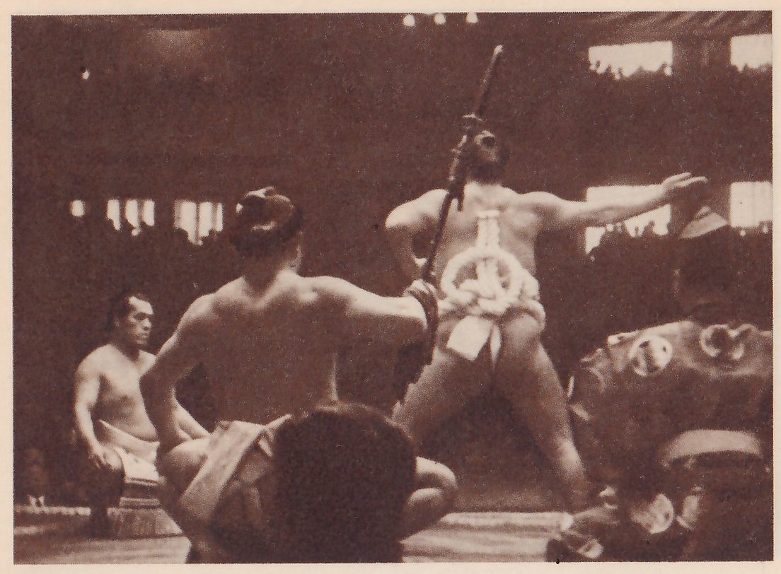 Dim photograph. At the center right a heavy man stands with his back to the camera, his legs apart, one arm stretched out to the right and his head tipped back, a thick rope ring with hanging strips at his back. In the left foreground another man, bare-backed with a topknot, sits turned away from the camera, and a long slender dark rod in his hand stands upright beside the standing man. Another man sits bare-chested at the far left, and a figure in a dark robe marked with round crests crouches at the right.