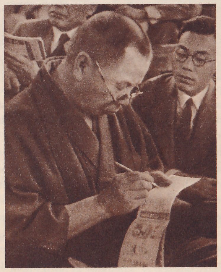 Close photograph of a man with a bald crown and round glasses, in a dark robe, leaning forward and writing with a pencil on a long strip of paper held in his other hand, the strip printed with small pictures and Japanese characters. At the right a man with round glasses, in a dark suit and tie, watches. Other spectators are partly seen behind them, one holding a printed booklet at the upper left.