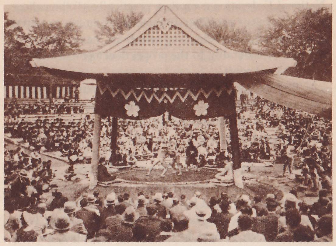 Photograph outdoors of an open pavilion with a wide curved roof and a gabled front with a lattice panel at its peak, held up by posts over a low round platform. A dark curtain with zigzag trim and two white blossom-shaped marks hangs under the front of the roof. Two men in light clothing stand locked together on the platform, with others seated around its edge. Tiers of seated spectators fill the ground around it, trees stand behind, an awning stretches out at the right, and the backs of the crowd, many in broad-brimmed hats, fill the foreground.