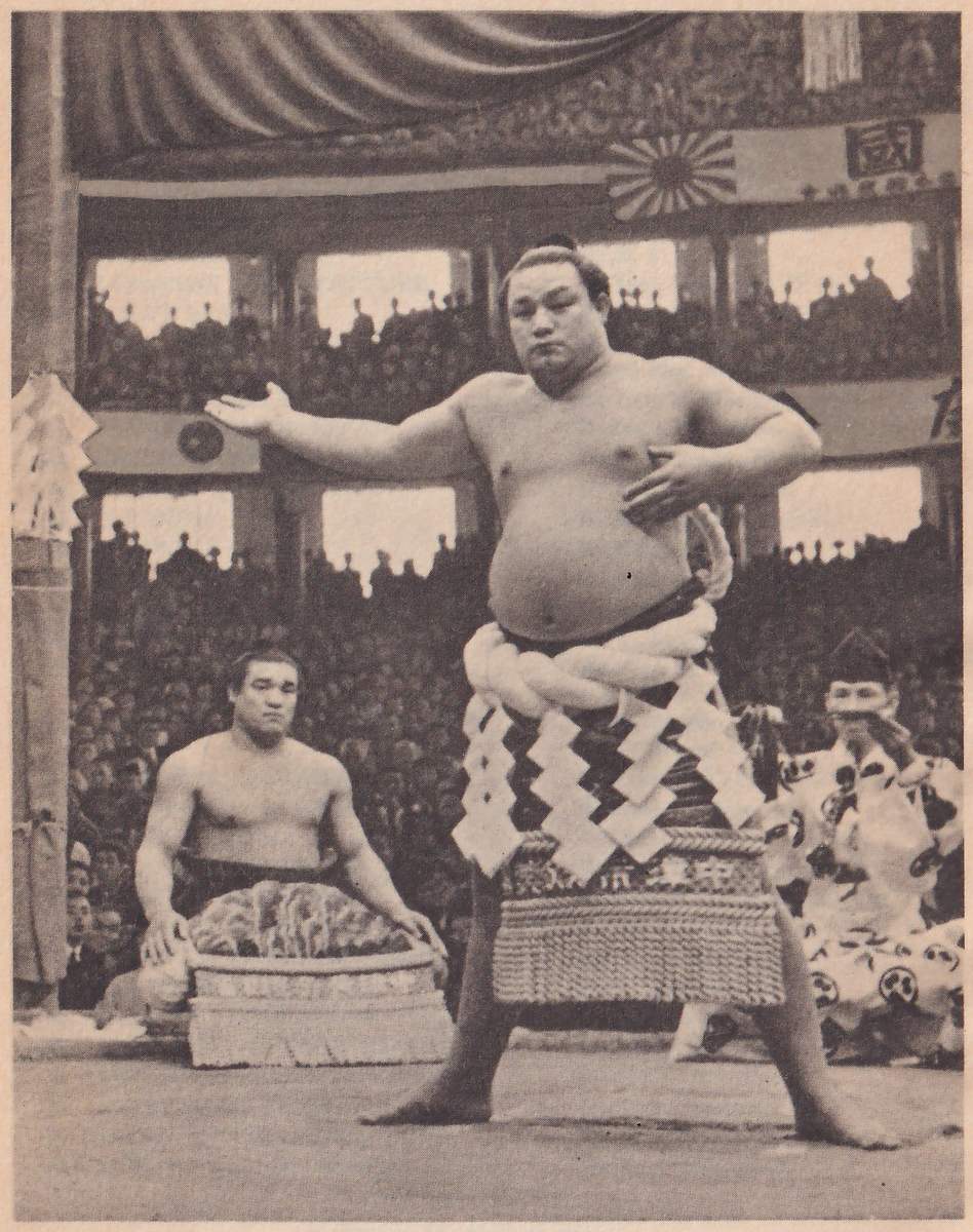 Photograph of a heavy, bare-chested man standing with his feet wide apart, one arm held out to the side with the palm open and the other bent with the hand at his waist. A thick twisted rope is wound around his middle over a belt, and an apron of heavy hanging cords with white zigzag paper strips stands out from the front of it. A topknot shows on his head. Behind him at the left a second bare-chested man sits, facing the camera, behind a wide round vessel, and at the right a man in a loose patterned robe and a tall black cap crouches. Tiers of spectators fill the background under a draped canopy at the top, with a flag showing a disc ringed by rays and a sign with one large Japanese character at the upper right.