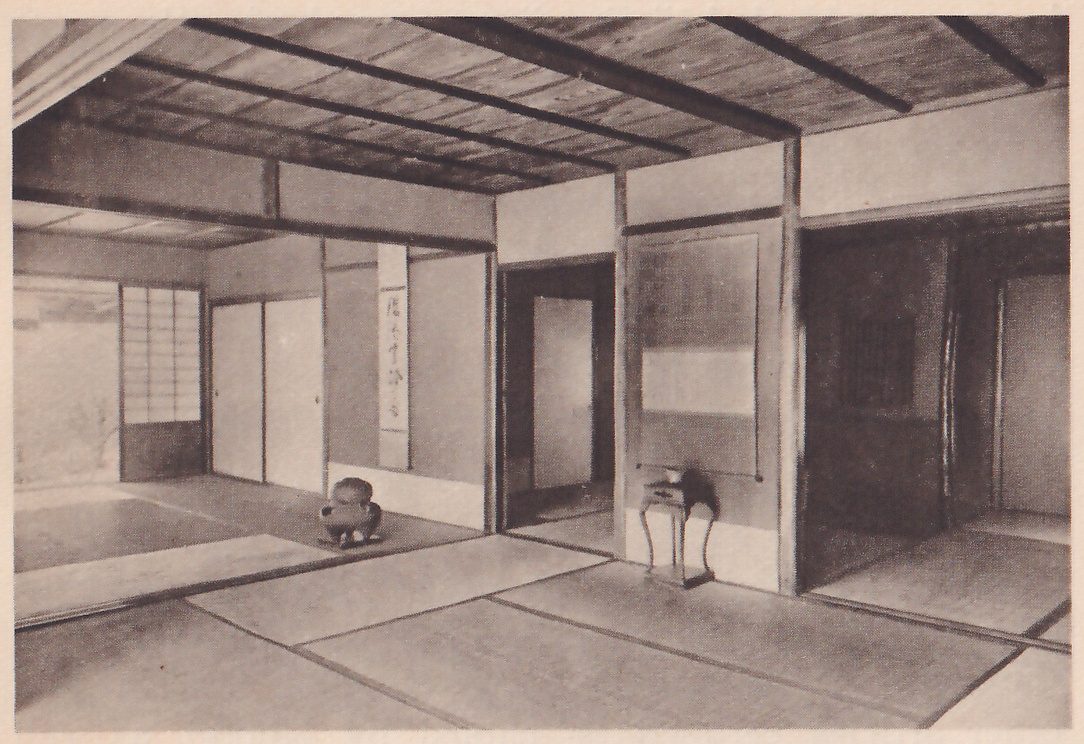 Photograph of an empty mat room with a ceiling of boards and dark beams: at the left a squat round dark vessel on the floor before a narrow hanging scroll, a window of paper panes beyond it; at the center a small stand on tall curved legs against a panel, and darker open rooms at the center and right.