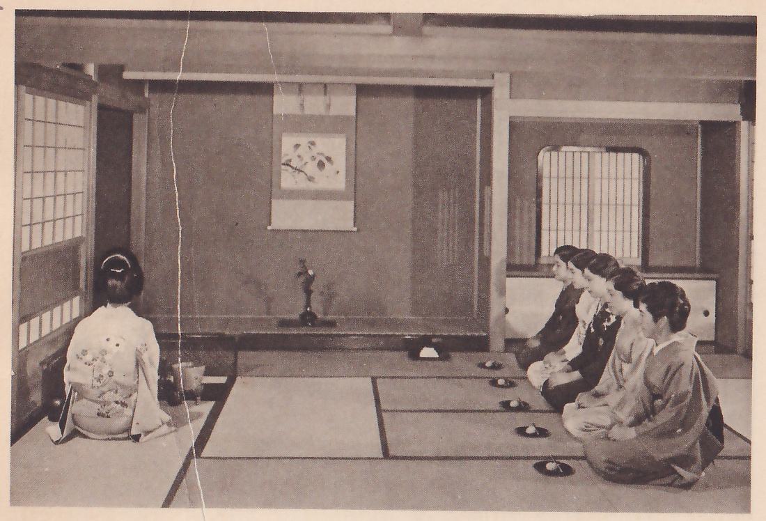 Photograph of a mat room with a hanging scroll of a flowering branch on the far wall and a small vase on the alcove floor below it: a woman in a light patterned kimono kneels at the left with her back to the camera, and a row of women in kimono kneel along the right side in profile facing left, with a row of small dark trays on the mats before them, each holding a white sweet.