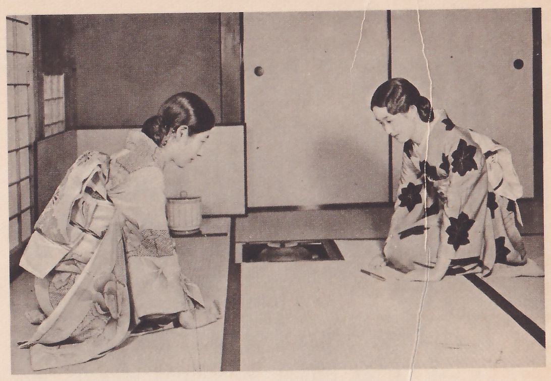 Photograph of two women in kimono kneeling on mats facing each other across a sunken hearth, both bowed forward: the woman at the left, in a pale kimono with a wide sash, faces right in profile; the woman at the right, in a kimono with large dark leaf-shaped patterns, faces left with her hands on the mat before her. A pale lidded jar stands behind the left woman and sliding doors fill the wall at the right.