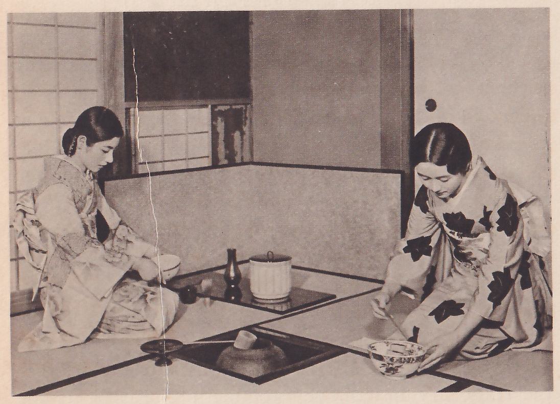 Photograph of two women kneeling in a mat room with a sunken hearth in the foreground: at the left a woman in a pale patterned kimono faces right with a pale bowl at her knee; at the right a woman in a kimono with large dark leaf-shaped patterns faces left, bending over a decorated bowl on the mat with a thin stick in one hand and the other hand on the bowl. Between them, on a dark surface, stand a lidded pale jar and a narrow dark vase, and in the hearth a dark iron pot.