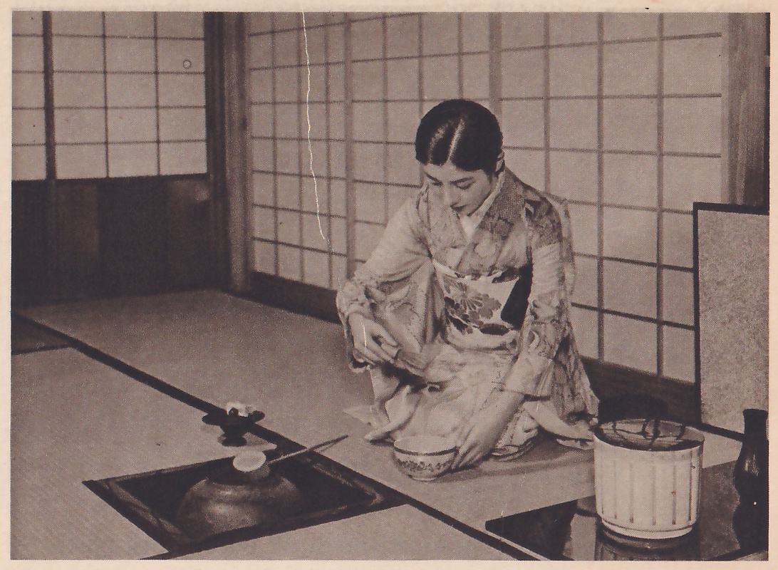 Photograph of one woman in a pale patterned kimono kneeling on mats before paper sliding doors, her head bowed, a small object held in one hand and a bowl on the mat in front of her. At the left a sunken hearth holds a dark iron pot with a thin stick across it; at the right stand a pale lidded jar with vertical lines and a dark vase.