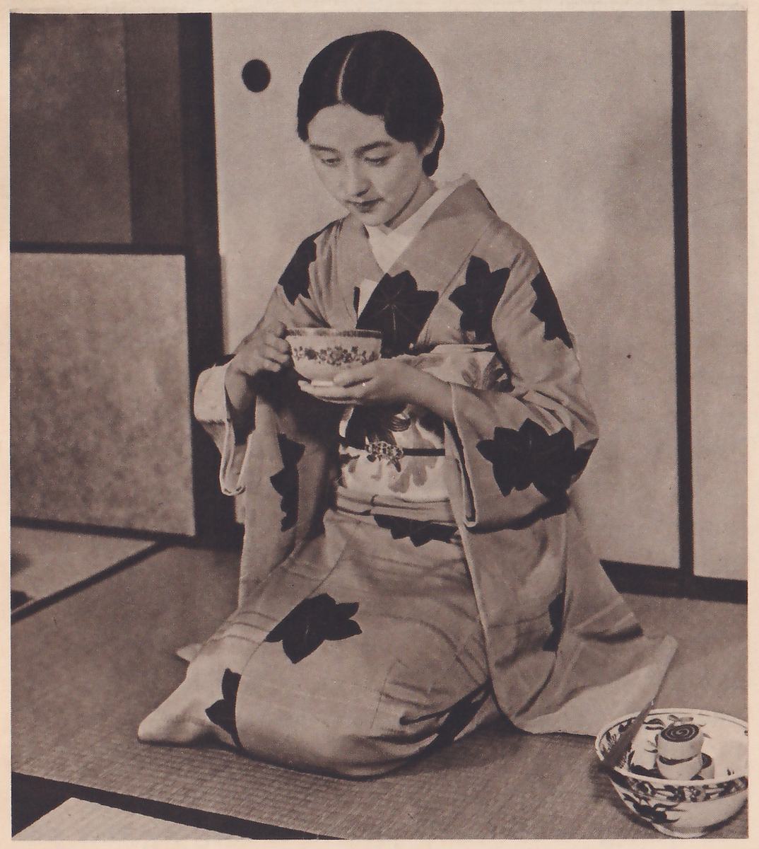 Photograph of a young woman in a kimono with large dark leaf shapes, kneeling on a mat before sliding panels and facing the camera, her head bowed toward a decorated cup on a saucer that she holds in both hands in front of her chest; at the lower right a decorated bowl on the mat holds a thin stick and a few small round objects.