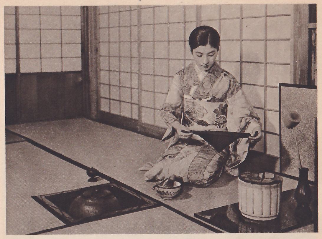 Photograph of a woman in a pale patterned kimono kneeling on mats before paper sliding doors, facing the camera with her head bowed, holding a dark triangular folded cloth stretched between her two hands at waist height. On the mat before her stands a small decorated bowl with a stick in it; at the left a sunken hearth holds a dark iron pot, and at the right a pale lidded jar with vertical lines stands on a dark surface beside a dark vase.