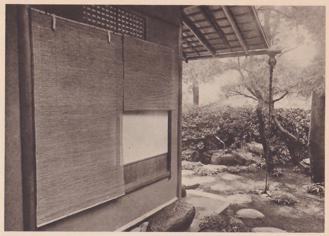 Photograph of the outer wall of a building at the left, covered by two woven reed screens hung at different heights, with a lattice panel above and a pale opening below them; at the right, under the eave of the roof from which a chain hangs to the ground, a garden of a pine tree, a clipped hedge, rocks and flat stepping stones, with a large rock in the foreground.