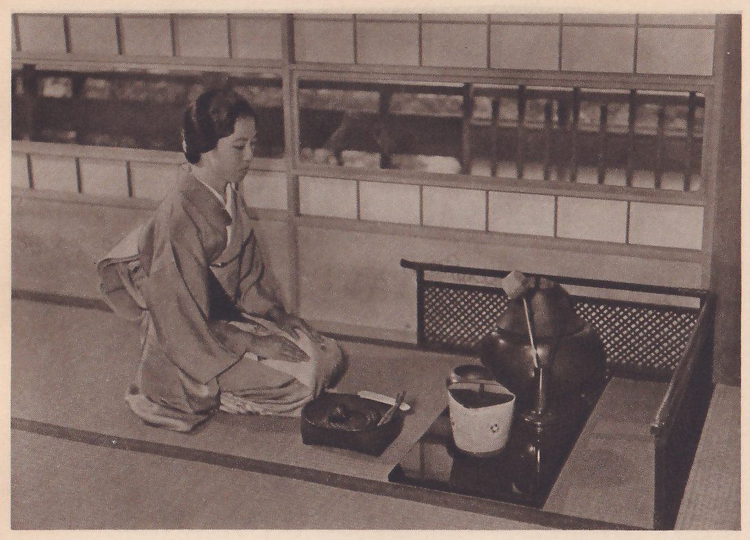 Photograph of a woman in a kimono with her hair up, kneeling in profile facing right on a mat before a wall of paper panels, her hands in her lap. In front of her stands a dark shallow box with small objects in it; at the right are a large dark round-bodied iron pot with a thin stick rising from it and a pale pail-shaped container with a small mark on it, on a dark glossy surface beside a low lattice screen.