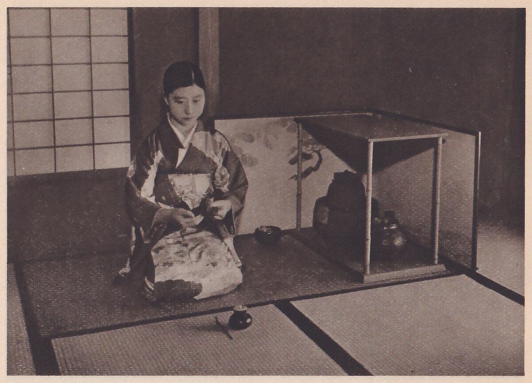 Photograph of a young woman in a dark patterned kimono kneeling on a raised mat platform, facing the camera with her head bowed and a small object in her hands; behind her at the right a square frame of thin posts with a flat top holds a tall dark jar and a smaller dark pot before a screen painted with branches, a small dark bowl sits on the platform beside her, and on the mat below it a small dark lidded pot has a thin stick lying beside it.