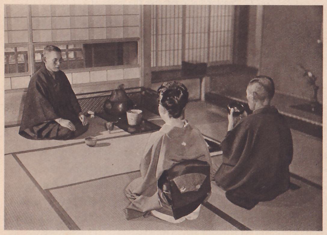 Photograph of three people in a mat room: at the left a man with round glasses in a dark kimono sits facing the camera with his hands on his knees; in the foreground a woman with her hair up, in a kimono with a dark sash, kneels with her back to the camera; at the right an older man in a dark kimono, seen from behind facing left, holds a dark bowl up in both hands near his face. Between them stand a large dark iron kettle and a pale pail-shaped container on a dark tray, and a slender flower arrangement is at the right edge.