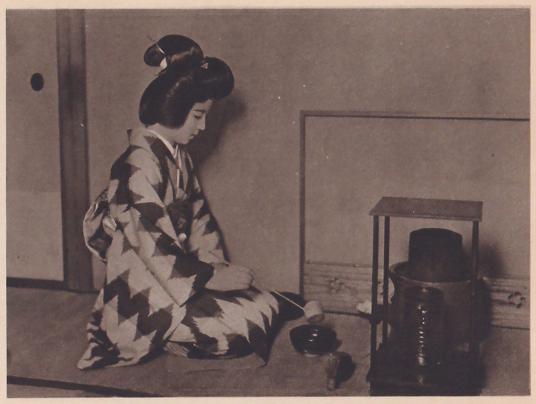 Photograph of a woman with her hair in a high bun, in a kimono with a bold zigzag pattern, kneeling in profile facing right on a mat, her hands in her lap holding the end of a thin-handled ladle whose cup is above a dark bowl on the mat in front of her. At the right a square stand of thin posts holds a large dark pot with a lid.
