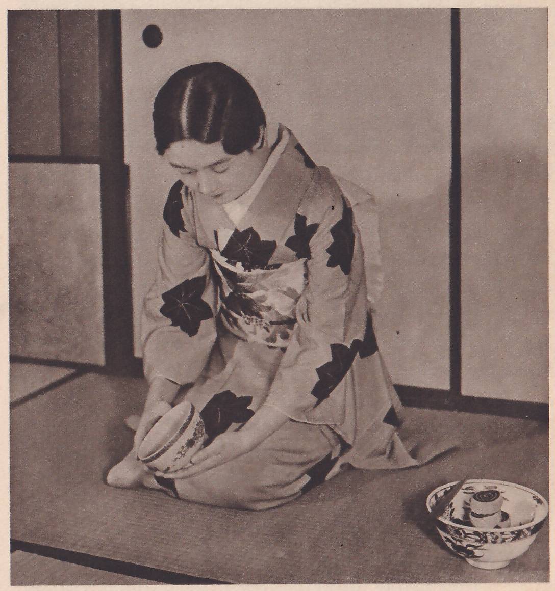 Photograph of a young woman in a kimono with large dark leaf shapes kneeling on a mat before sliding panels, facing the camera with her head bowed, holding a decorated bowl in both hands low in front of her knees; at the lower right a decorated bowl on the mat holds a thin stick and a few small round objects.