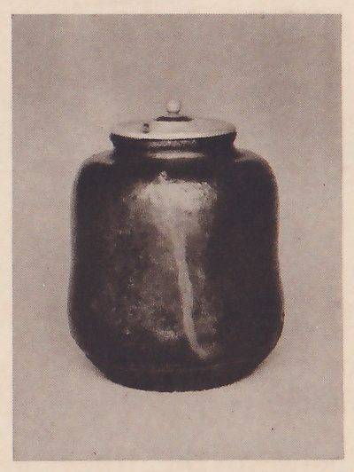 Photograph of a dark glazed jar on a plain gray ground, with a pale lid topped by a small knob and a pale streak of glaze running down its front.