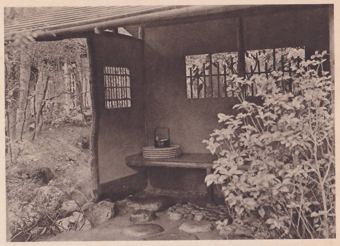 Photograph of an open-sided garden shelter under a sloping roof: a low wooden bench against a plain plastered wall holds a stack of flat round mats and a small box with a handle, with a small lattice window in the wall at the left and a wider one at the right. Trees stand at the left, a leafy shrub at the right, and rocks and flat stepping stones lie in front.