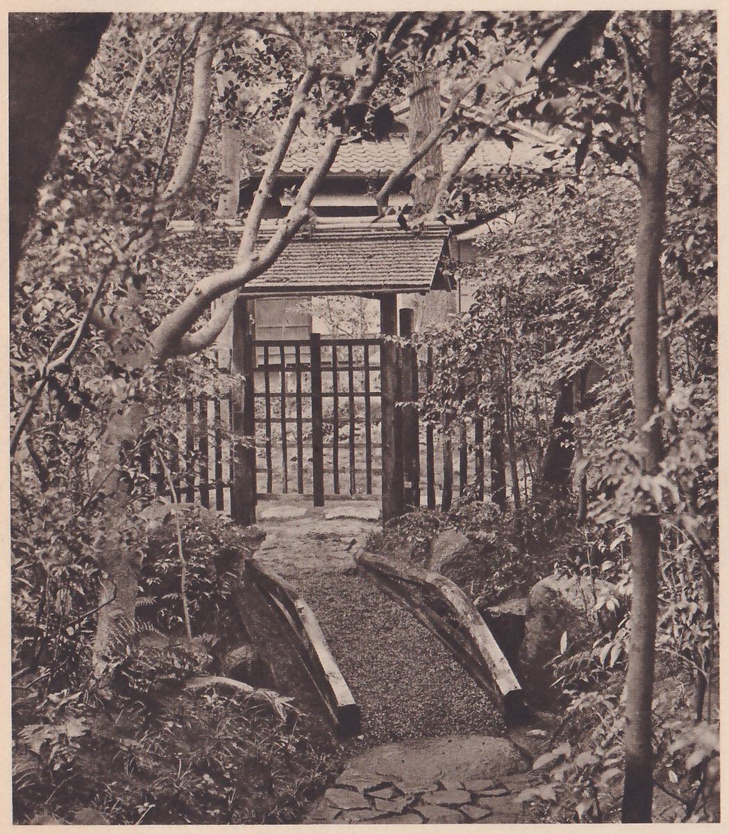 Photograph of a garden path of fine gravel running between two slanted plank edges toward a gate of upright dark wooden bars under a small shingled roof, with a larger tiled roof behind it; trees and shrubs frame the view, a slim trunk at the right and a bent branch crossing from the left, and flat stones lie at the foot of the path.