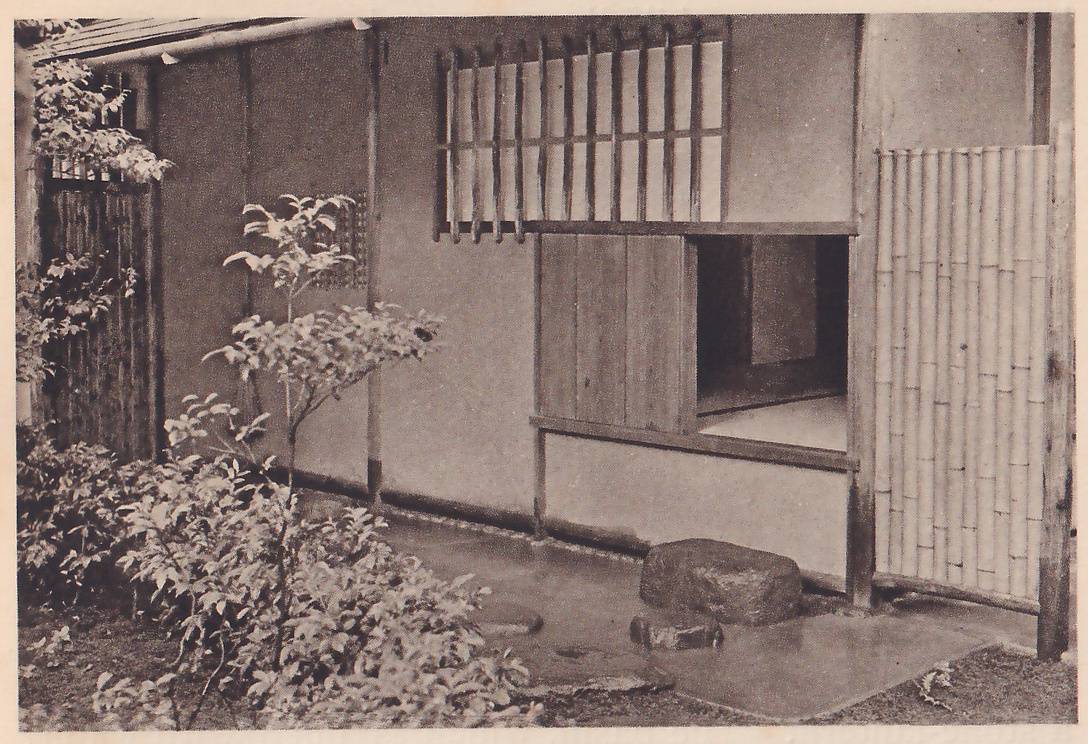 Photograph of the plastered outer wall of a small building: a low square opening at the center right with a wooden board door slid open on a dark room and a pale floor, a window of vertical wooden bars over paper above it, a stone and a flat stepping stone before it, a fence panel of upright bamboo poles at the right and a small leafy shrub at the left.