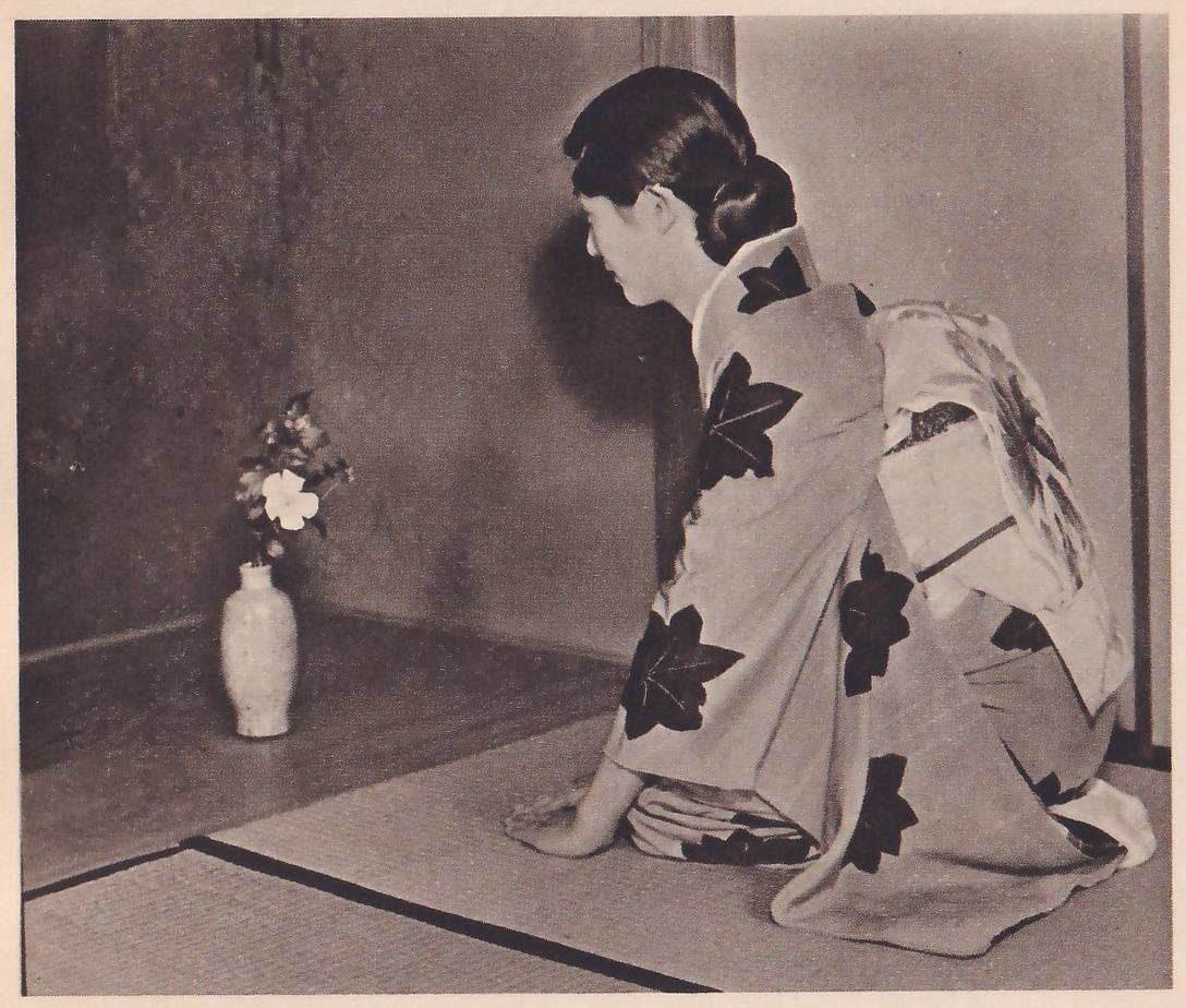 Photograph of a young woman in a kimono with large dark leaf shapes, kneeling at the right seen from behind and leaning forward with one hand on the mat, her face turned left toward a small pale vase on the floor of an alcove at the left, which holds a few flowers, one of them white.