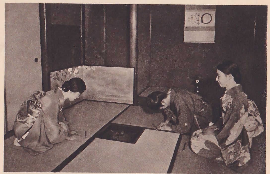 Photograph of three women in kimono kneeling in a mat room around a square sunken hearth: the one at the left bows forward facing right, the one at the center bows with her head near the mat facing left, and the one at the right kneels upright facing left. At the back a low screen painted with flowers stands at the left, and a hanging scroll showing a large circle hangs at the upper right.