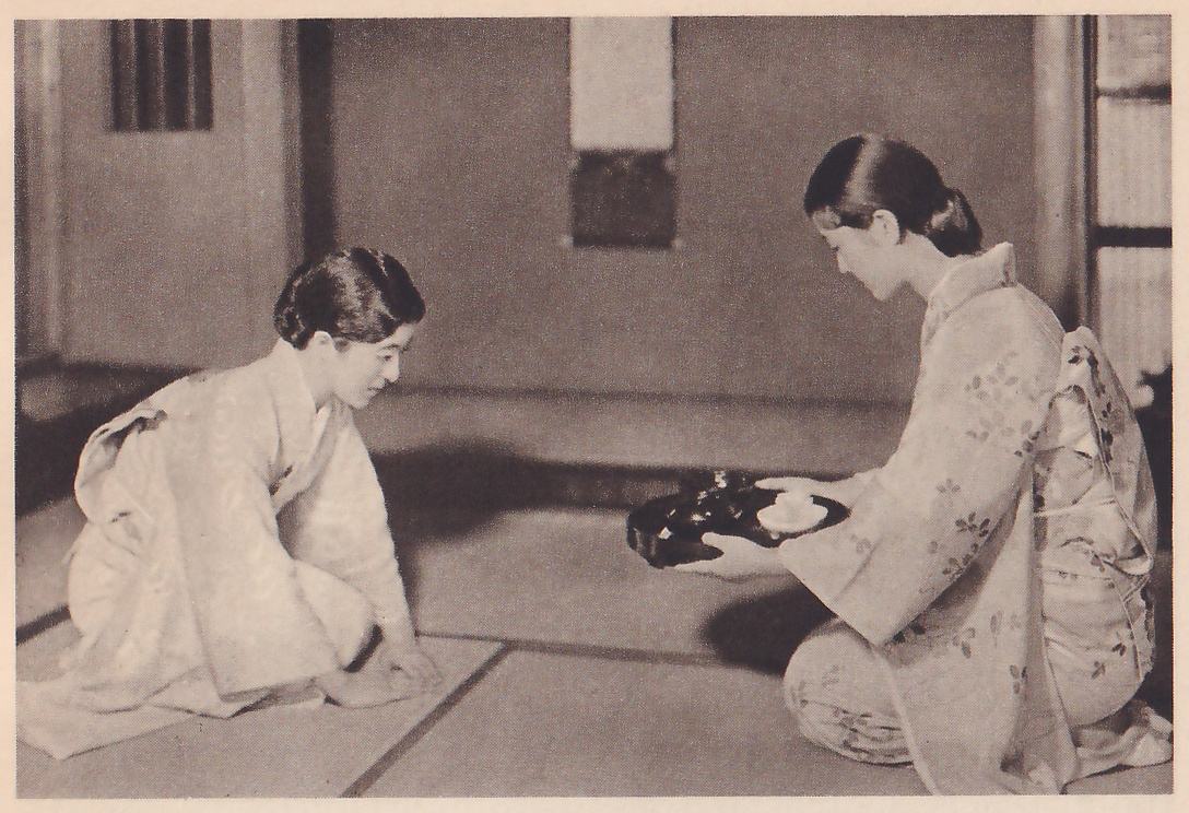 Photograph of two women in kimono kneeling in a mat room: the one at the left bows forward facing right, her hands on the mat; the one at the right, in profile facing left, holds out a dark tray with a small white cup on it in both hands. A hanging scroll is on the wall behind them.