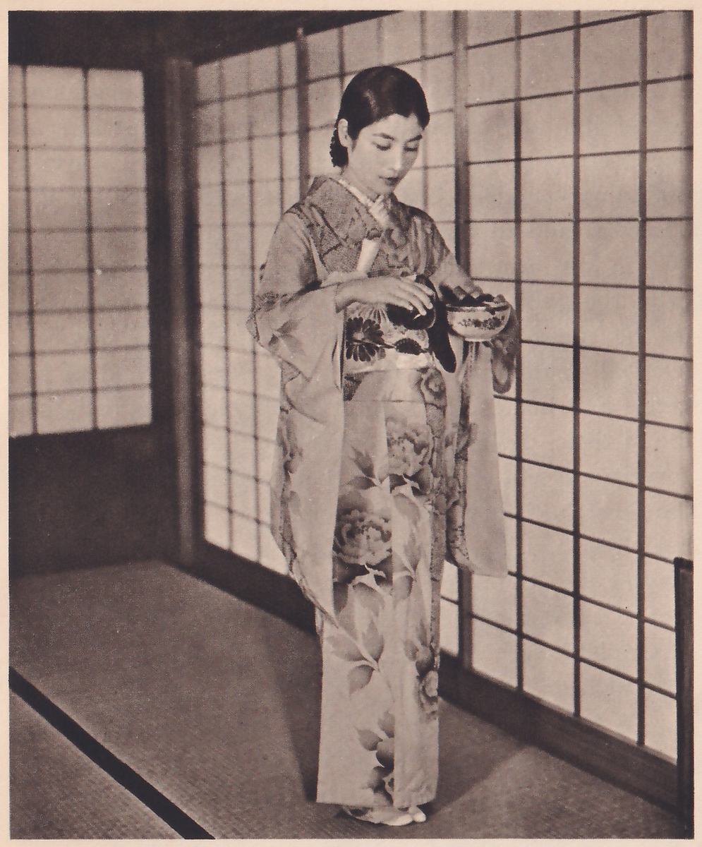 Photograph of a woman standing in a mat-floored room before paper sliding doors, in a long kimono with large flower patterns, her head bowed, holding a decorated bowl in one hand and a dark rounded object in the other at waist height.