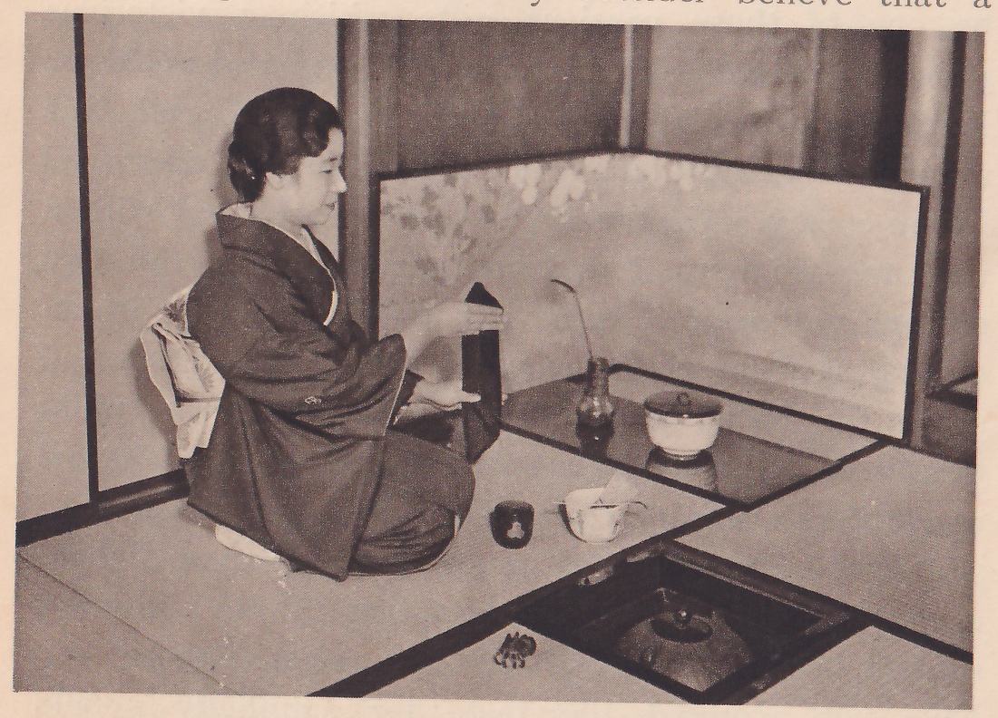 Photograph of a woman in a dark kimono kneeling in profile facing right on a mat, holding a dark folded cloth upright between her hands in front of her; on the mat before her are a small round dark lidded pot and a pale bowl with a stick in it. At the back a low folding screen painted with pale flowers stands in the corner, and on a dark board before it stand a small vase with one slender stem and a pale lidded bowl; a sunken hearth with a dark lidded pot is at the lower right.