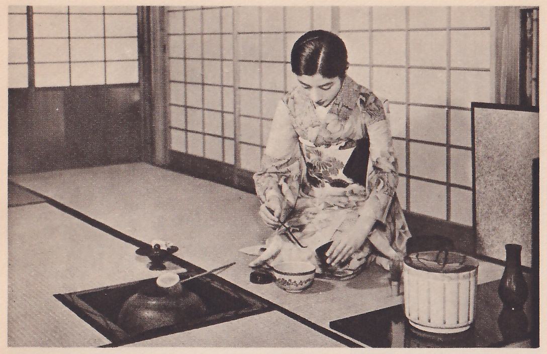 Photograph of a woman in a pale patterned kimono kneeling on mats before paper sliding doors, facing the camera with her head bowed, holding a thin stick in one hand above a patterned bowl on the mat in front of her. At the left a sunken hearth holds a dark iron pot with a pale lid and a long thin handle laid across it; at the right a white lidded jar with vertical lines and a dark vase stand on a dark surface beside a screen.